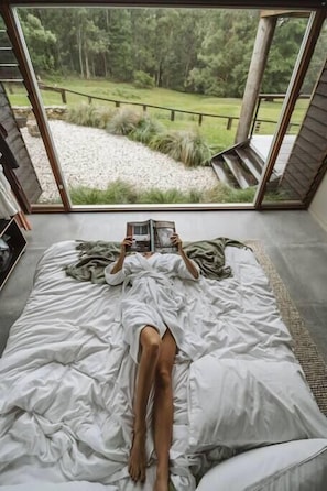 A person lying on a bed with a book, in a room with a large window overlooking a garden.