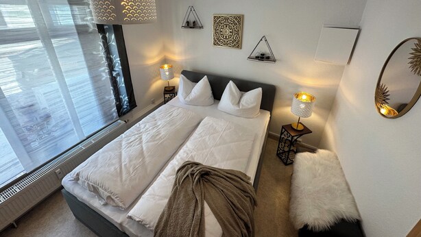Una camera da letto moderna con un letto grande, due lampade ai comodini, uno specchio decorativo e una finestra con persiane.