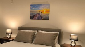 Un lit bien fait, avec une serviette pliée, deux tables de chevet avec des lampes et un tableau représentant un coucher de soleil accroché au mur.