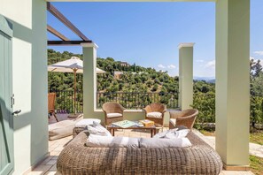 Ein Balkon mit Korbmöbeln, einem Tisch mit einem Buch und Blick auf eine hügelige Landschaft.