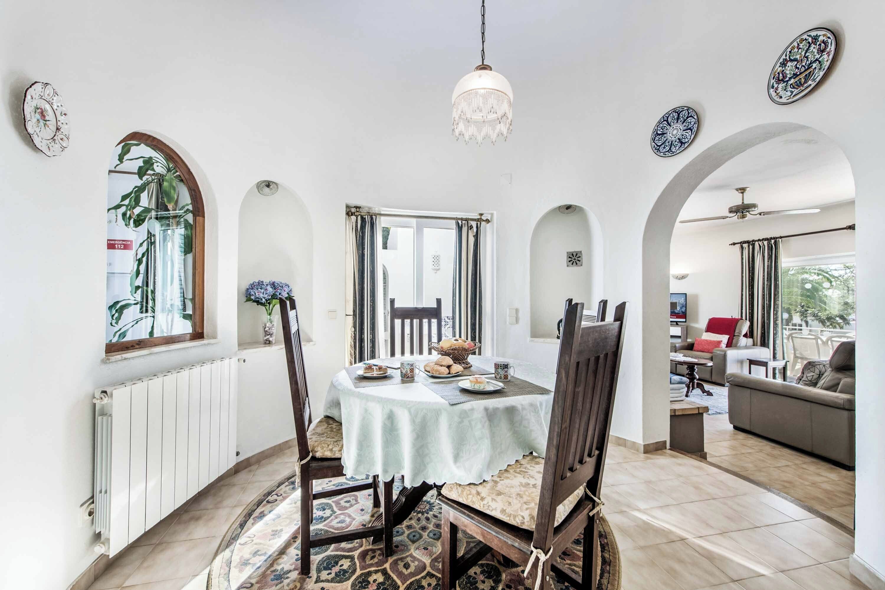 A dining area with a round table, chairs, and a chandelier. There are decorative plates on the walls and a window with curtains.