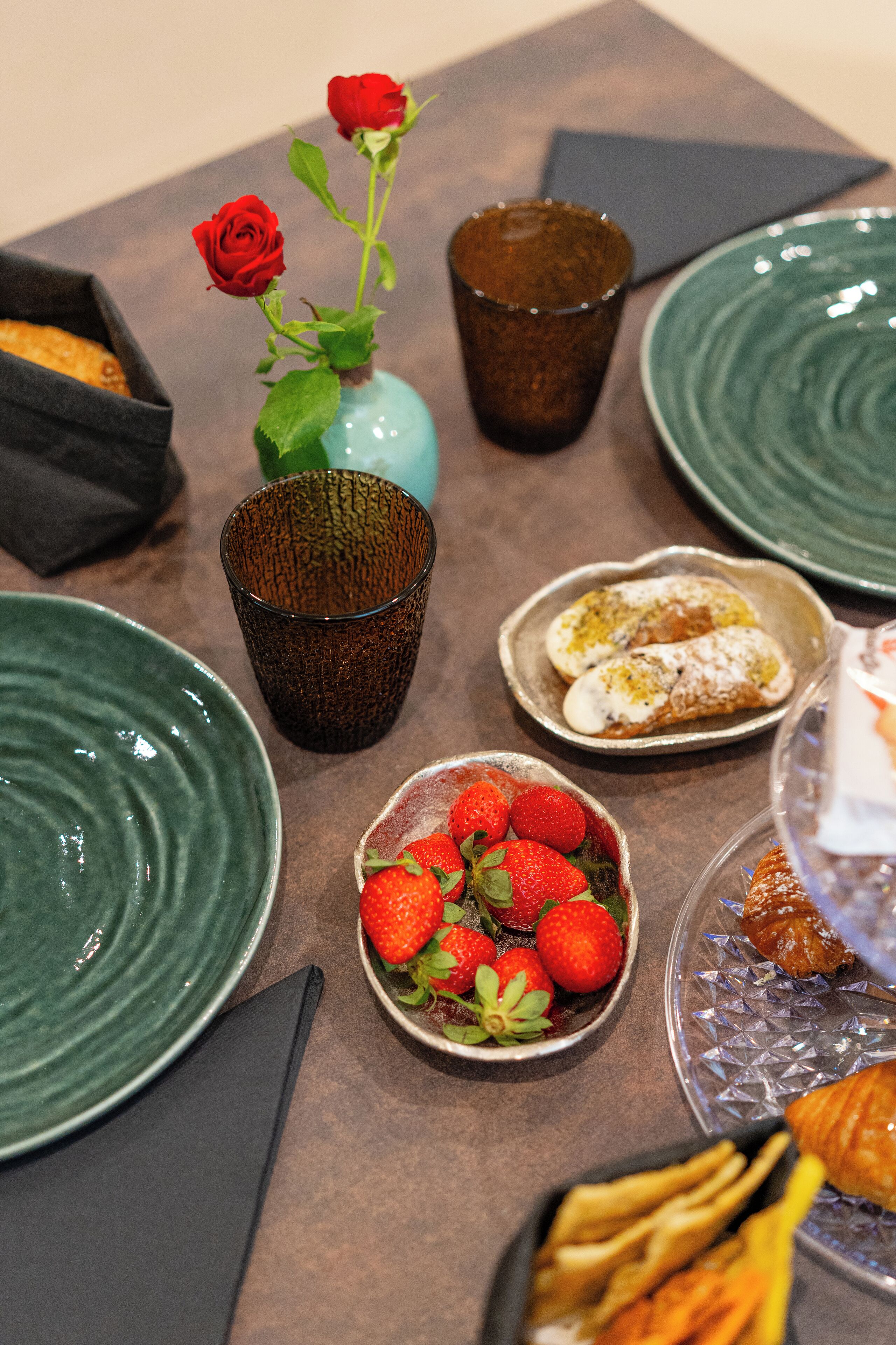 A table with various dishes, including a bowl of strawberries, a plate of pastries, and a vase with a rose.