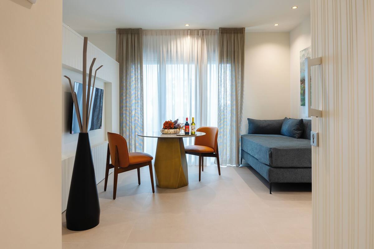 A modern living room with a round table, two chairs, a sofa, and a large window with curtains.