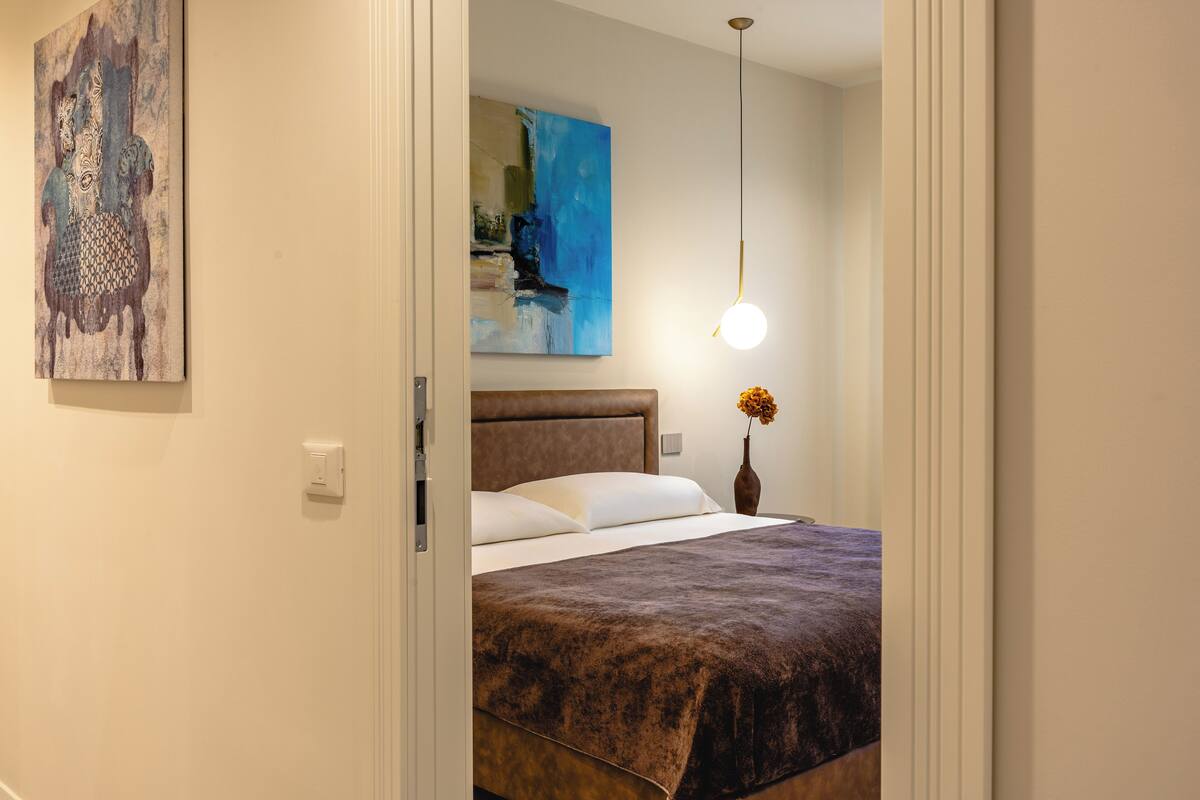 A bedroom with a bed, a painting, a vase with flowers, and a hanging light fixture.
