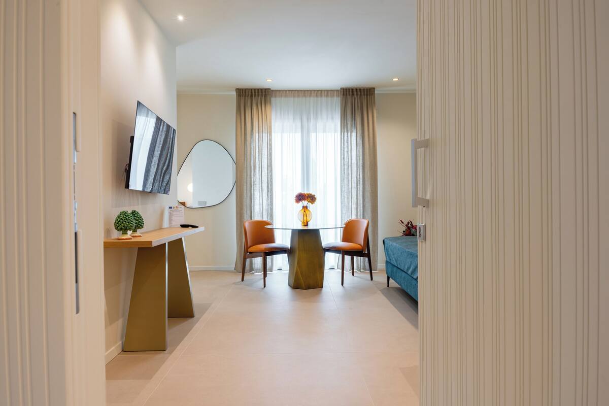 A modern hotel room with a dining area, a TV mounted on the wall, a round table with chairs, and a bed with a blue cover.