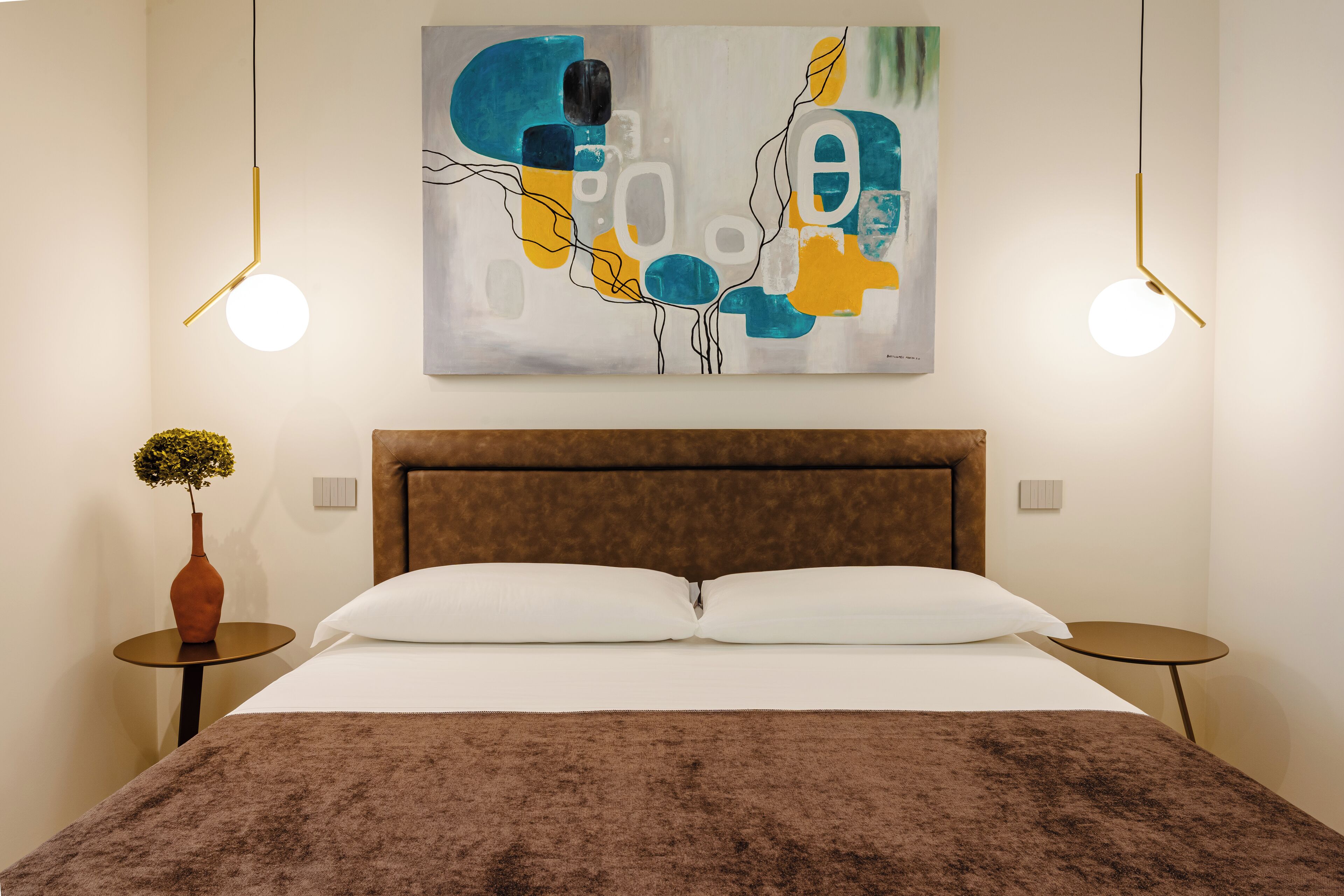 A modern hotel room with a large bed, two bedside tables, a painting on the wall, and two pendant lights.
