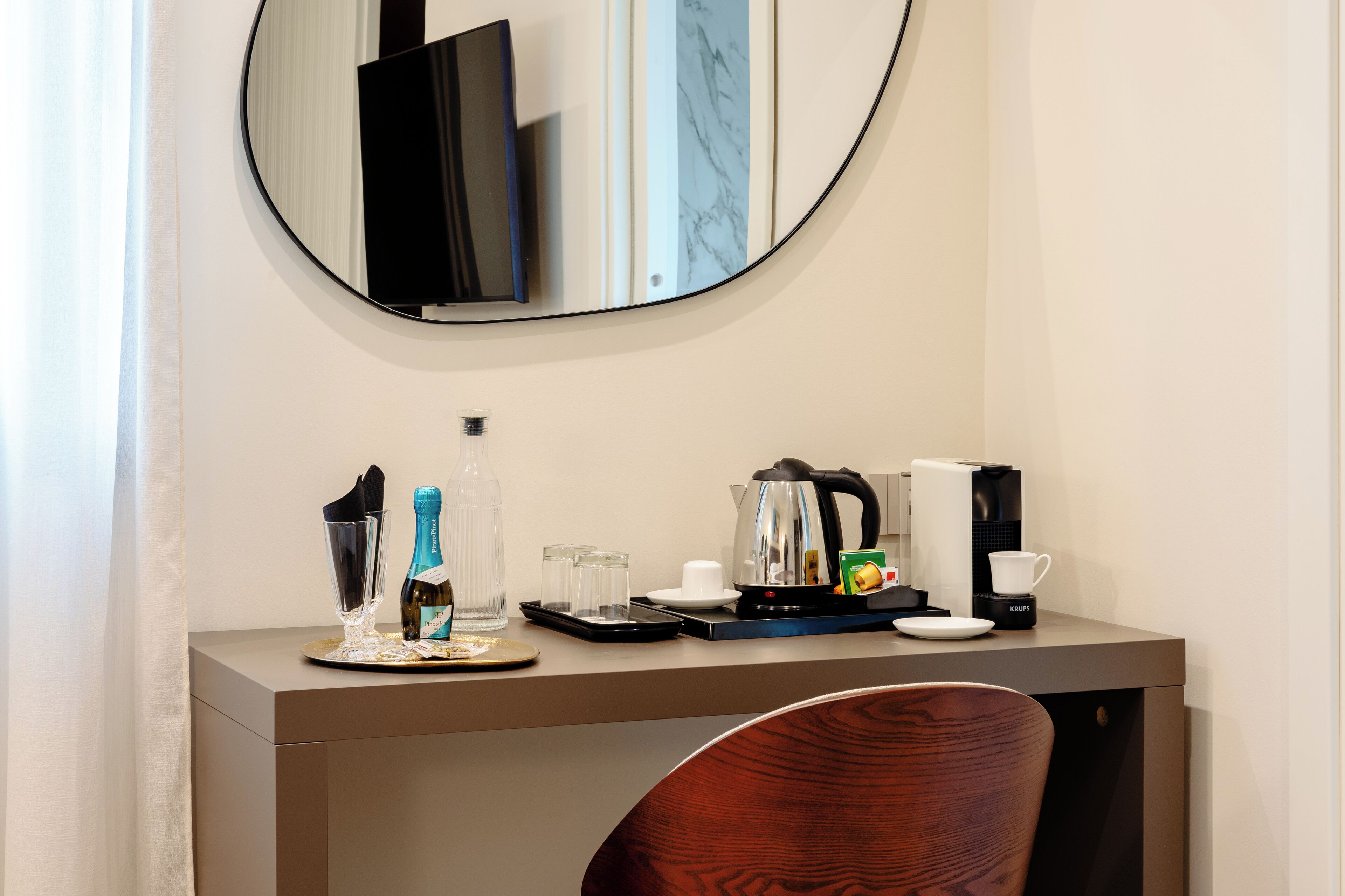 A hotel room with a wooden desk, a mirror, a coffee maker, a kettle, a bottle of champagne, and a cup.