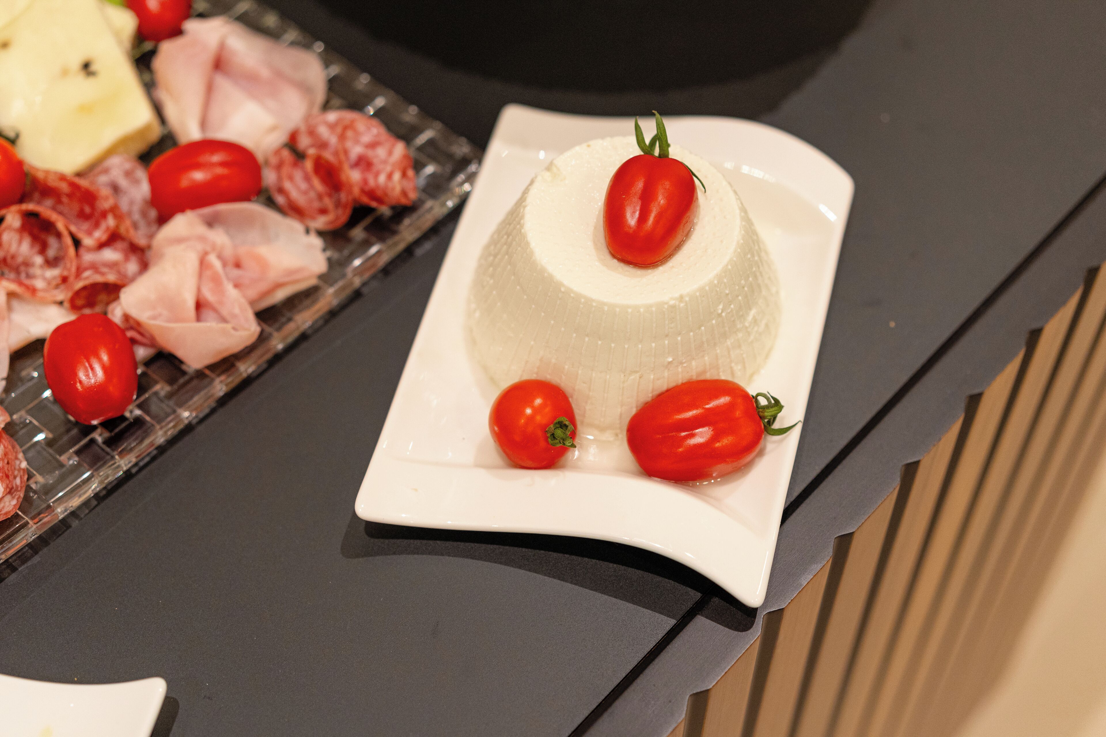 Une assiette blanche sur laquelle repose un plat en forme de dôme, agrémenté de tomates cerises et d’une feuille verte, le tout posé sur un plan de travail noir.