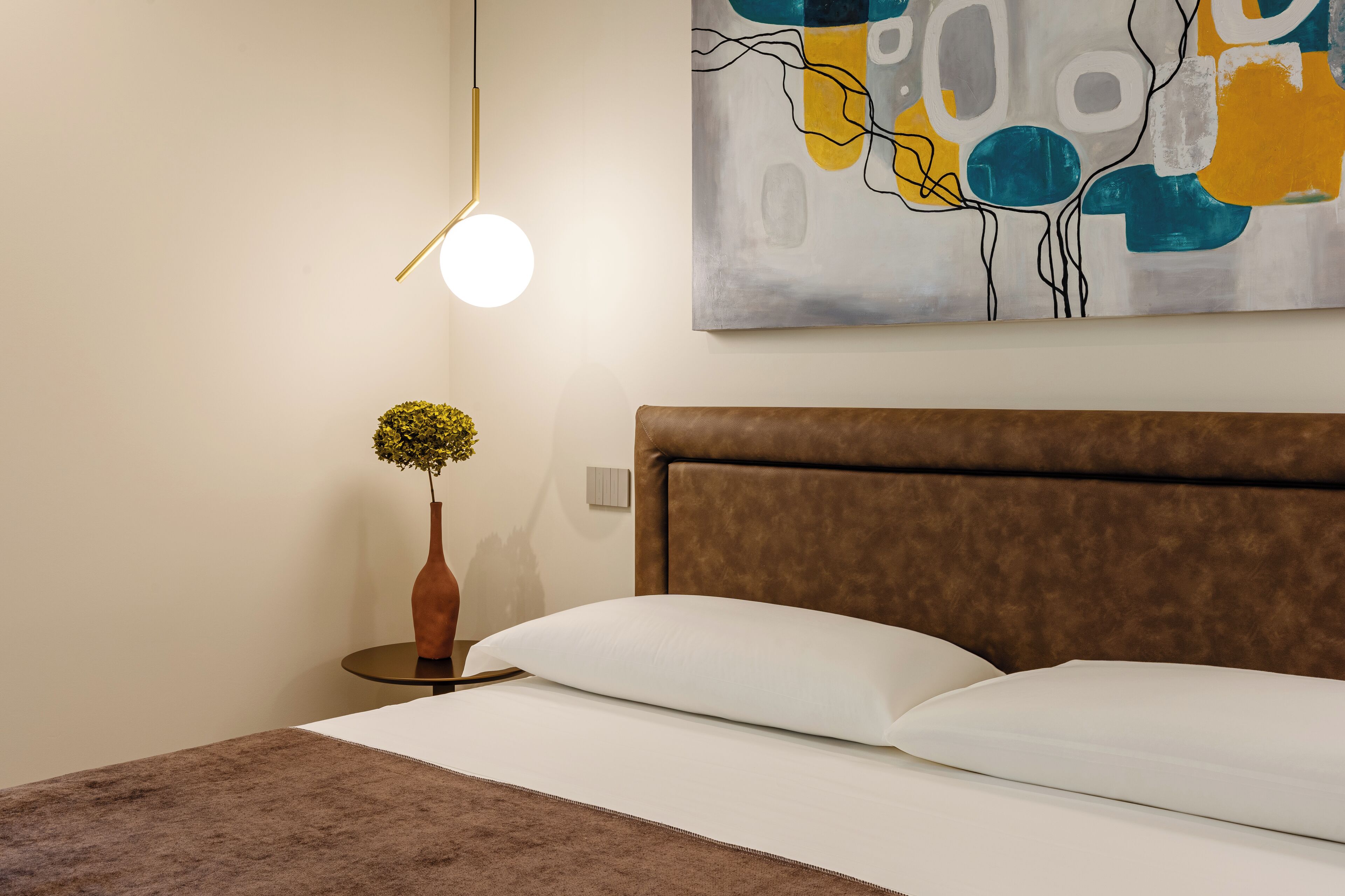 A neatly made bed with a brown headboard, a round table with a vase of flowers, and a modern abstract painting on the wall.