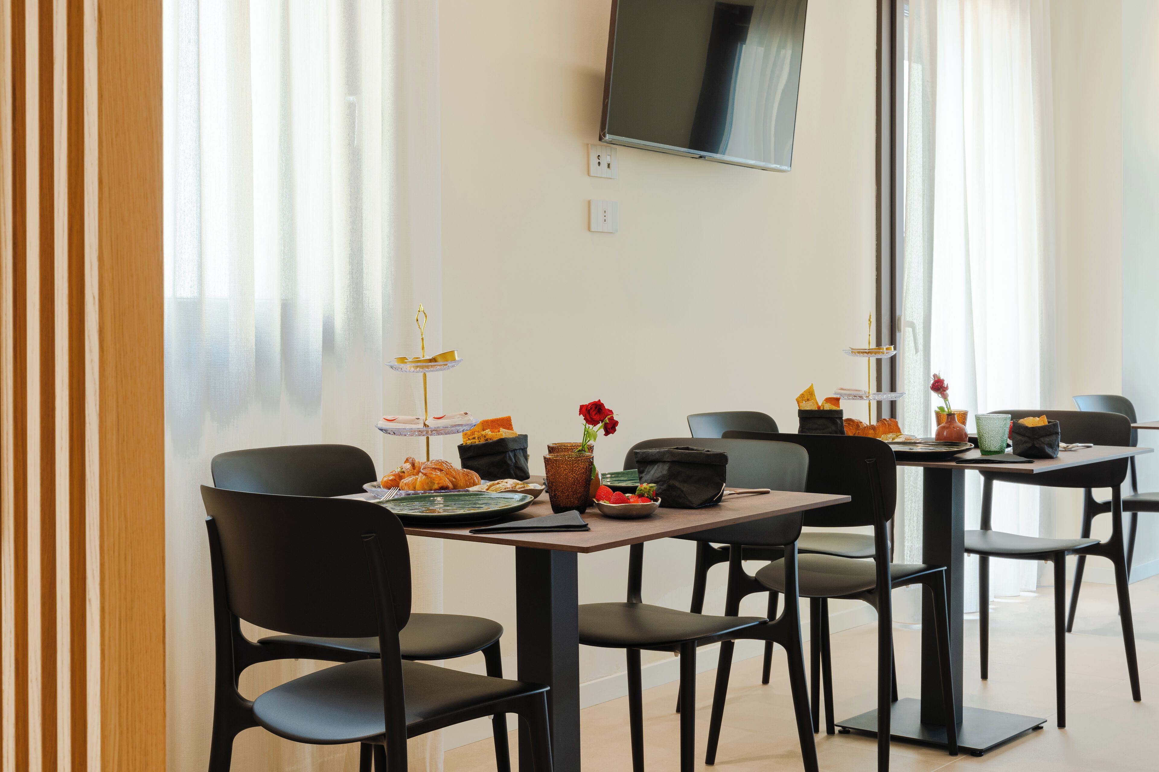 A modern dining area with black chairs and tables, a flat-screen TV mounted on the wall, and a window with sheer curtains.