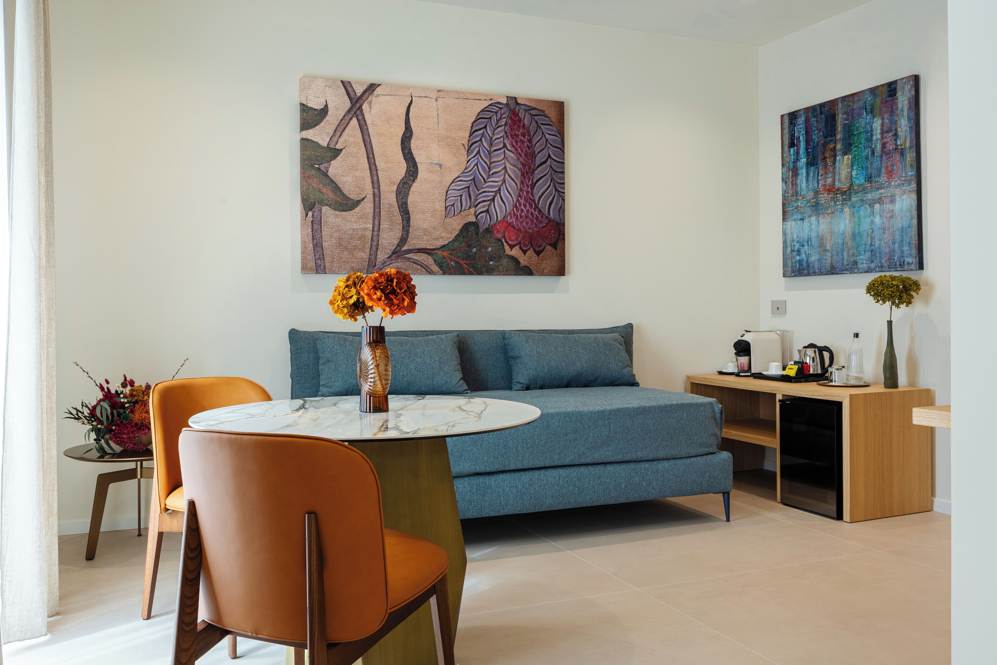 Una sala de estar moderna con un sofá azul, sillones naranja, una mesa de mármol y dos obras de arte en las paredes.