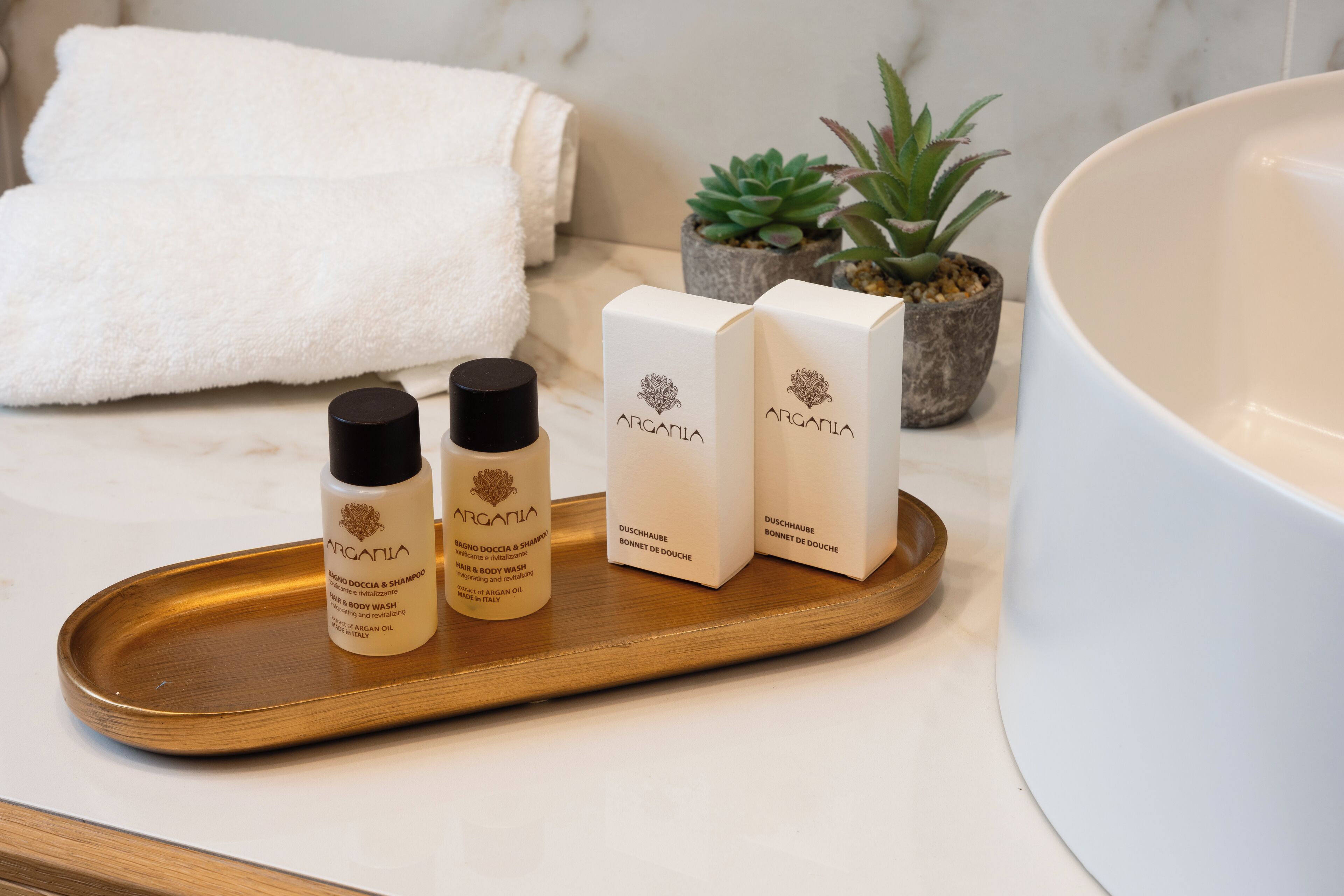 Un mobile bagno con un lavabo bianco, un vassoio di legno con tre flaconi di bagnoschiuma Argania e una piccola pianta in vaso.