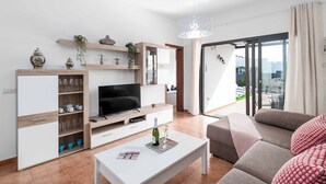 Un salon moderne doté d’un téléviseur à écran plat, d’étagères en bois ornées d’objets décoratifs, d’une table basse blanche sur laquelle repose un magazine, et d’une grande fenêtre offrant une vue sur la ville.