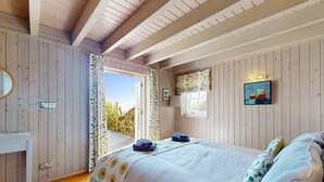 A bedroom with wooden walls, a bed with a floral bedspread, a bedside table, a mirror, and a window with curtains. - Talarfor - Two Bedroom Cottage, Sleeps 4 (Rhiw)