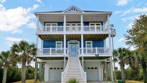 A two-story beachfront house with a balcony on each floor, a front garage, and a staircase leading to the entrance. - Luxurious Escape Steps from the Beach w/ Pool, Dock, & Stunning Views (North Topsail Beach)