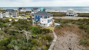 A coastal house with a large deck and a swimming pool, surrounded by dense vegetation and a view of the ocean. - Luxurious Escape Steps from the Beach w/ Pool, Dock, & Stunning Views (North Topsail Beach)