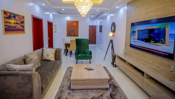 A modern living room with a grey sofa, a green armchair, a wooden coffee table, and a TV on a wooden stand. - Novabase Apartment (Lekki)