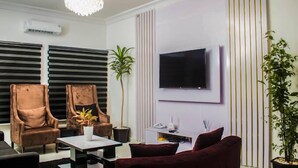 A modern living room with a flat-screen TV mounted on the wall, two leather armchairs, a coffee table, and a chandelier. - Novabase Apartment (Lekki)
