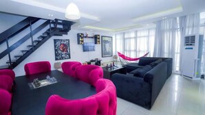 A modern living room with a black sofa, bright pink chairs, a dining table, and a staircase. - Novabase Apartment (Lekki)