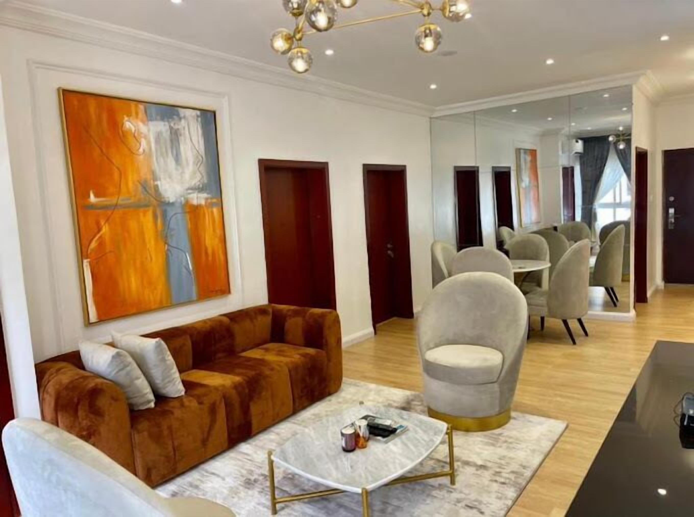 A modern living room with a brown velvet sofa, a white coffee table, and a large abstract painting. There is a dining area with grey upholstered chairs and a chandelier.