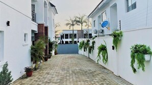 A narrow alleyway with a paved walkway flanked by white buildings, some with satellite dishes and potted plants. - Novabase Apartment (Lekki)