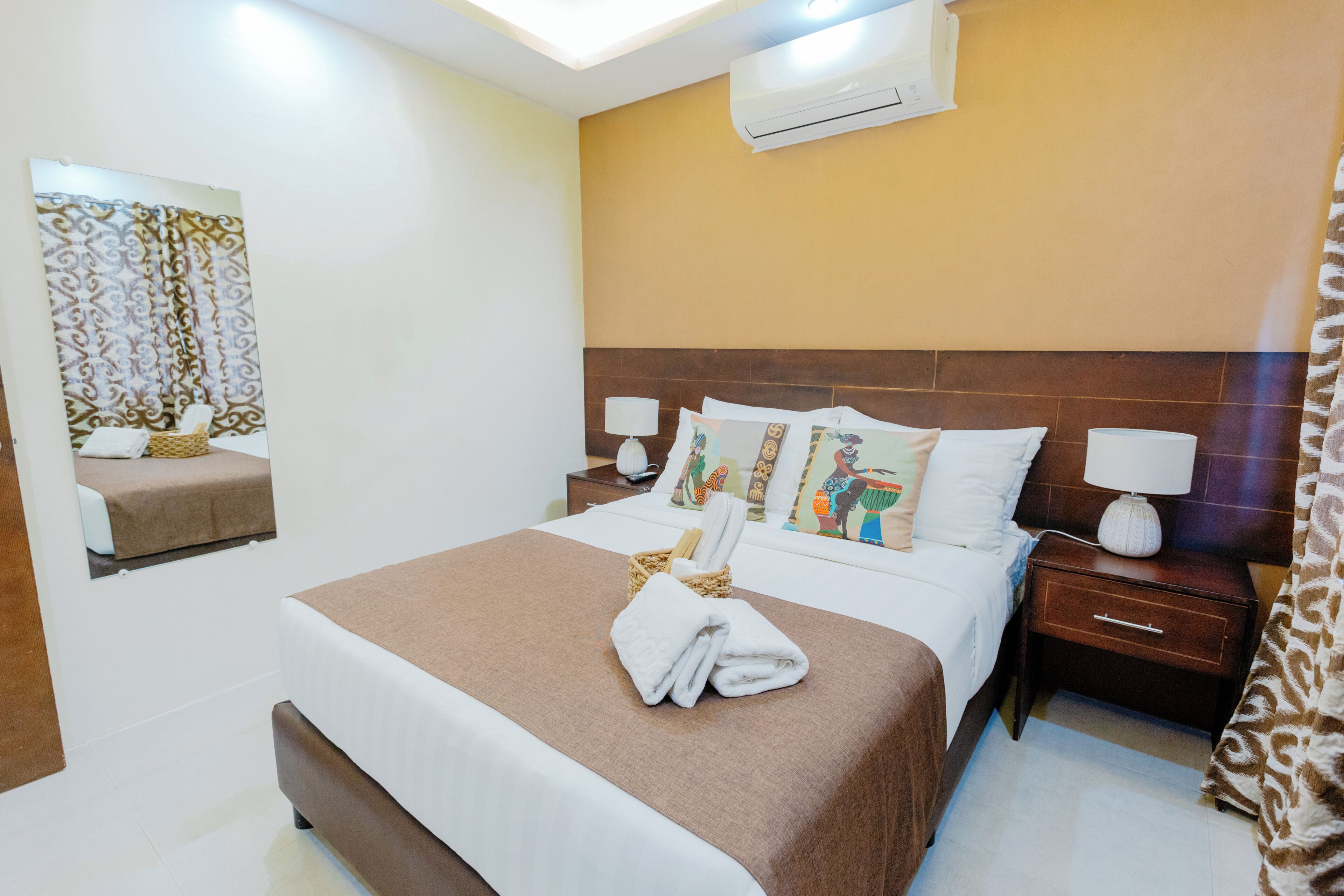 Habitación de hotel con cama, mesita de noche, aire acondicionado de pared y espejo con marco decorativo.