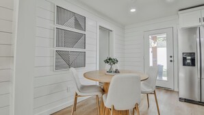 A modern kitchen with white cabinetry, a stainless steel refrigerator, a round wooden table with two white chairs, and decorative wall art. - The Pops-Stylish newly remodeled 3-bedroom house with indoor/outdoor living area (Lubbock)