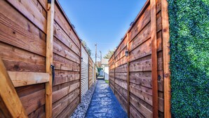 A narrow wooden corridor with a stone pathway, flanked by wooden walls and a green hedge on one side. - The Pops-Stylish newly remodeled 3-bedroom house with indoor/outdoor living area (Lubbock)