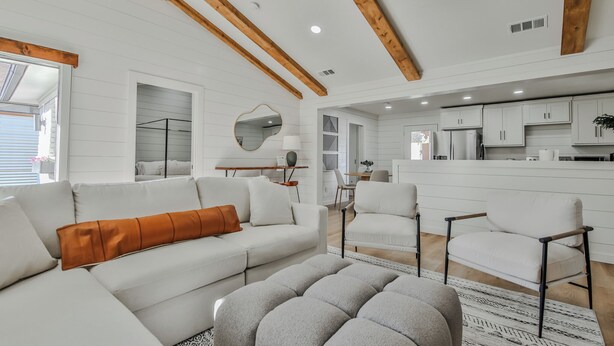 A spacious living area with a sofa, ottoman, and chairs, wooden beams on the ceiling, and a kitchen in the background. - The Pops-Stylish newly remodeled 3-bedroom house with indoor/outdoor living area (Lubbock)
