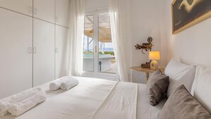 A hotel room with a bed, white curtains, a view through a glass door, a nightstand with a lamp, and a painting on the wall. - White Cycladic House With Jacuzzi (Hersonissos)