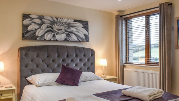 A bedroom with a tufted headboard, a large floral painting, a bed with white linens and a purple pillow, two bedside tables with lamps, and a window with blinds. - 1 bedroom accommodation in Carnforth (Carnforth)