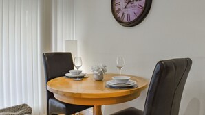 A dining area with a round wooden table, two chairs, a wall clock, and a window with blinds. - 1 bedroom accommodation in Carnforth (Carnforth)