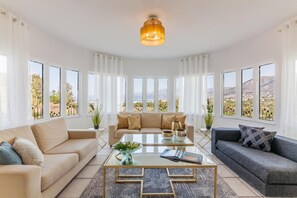 Ein geräumiges Wohnzimmer mit einem beigen Sofa, einem grauen Sofa und einem Couchtisch. Große Fenster bieten Blick auf eine gebirgige Landschaft.