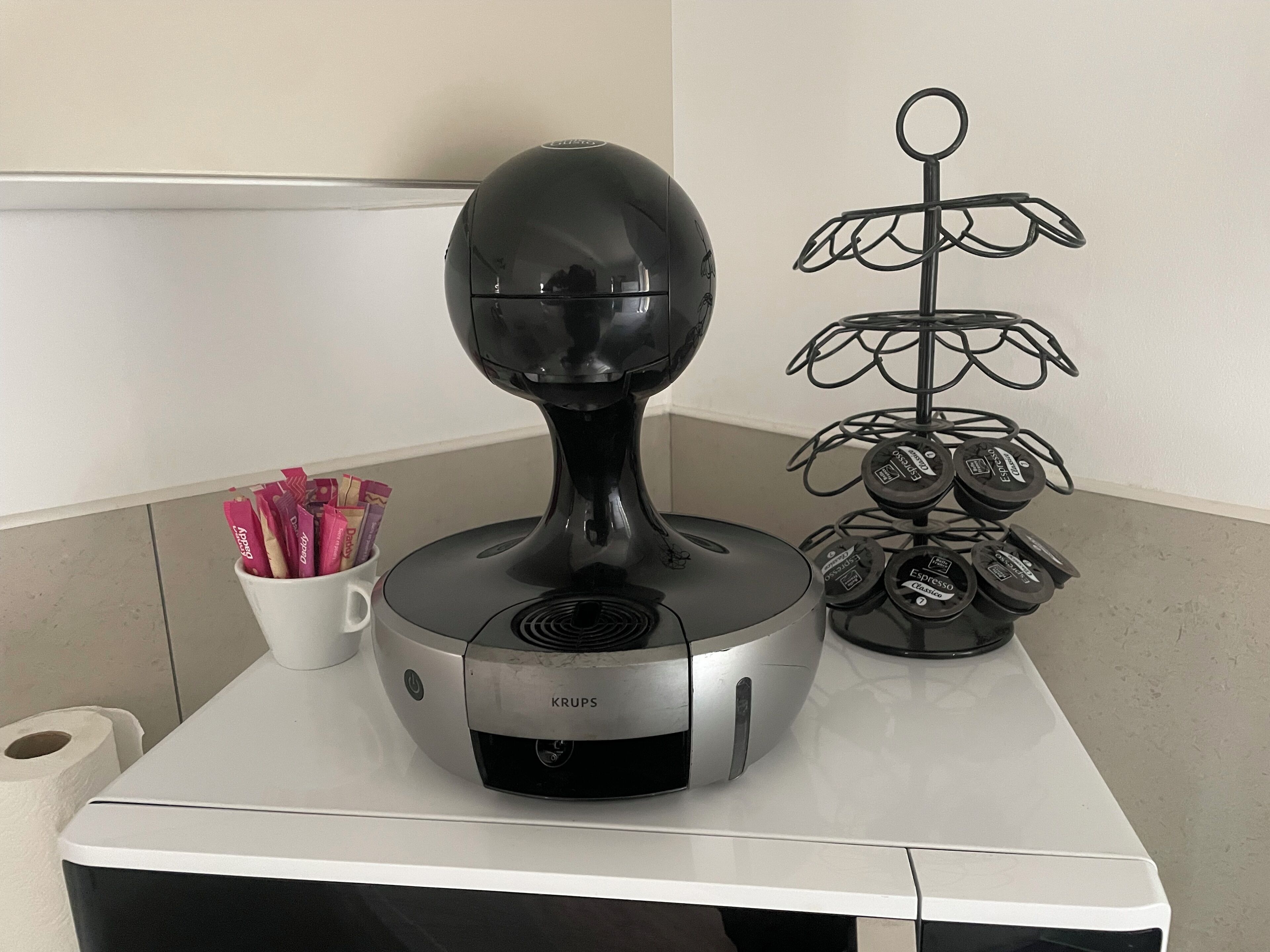 A Krups coffee machine on a white countertop with a coffee pod holder and a metal stand with glasses.