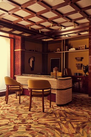 Une pièce avec un tapis à motifs, un bureau en bois avec deux chaises, un radiateur et une étagère contenant divers objets.