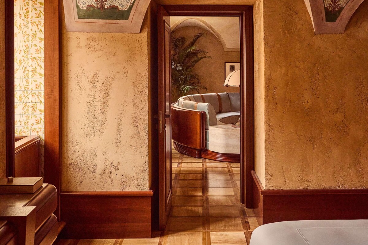 Un pasillo con un banco de madera, una puerta que da a una habitación con un sofá y una planta, y una pared con diseño.