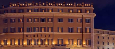 A multi-story hotel with a prominent 'MINERVA' sign, a central entrance with a canopy, and a series of balconies with lights.
