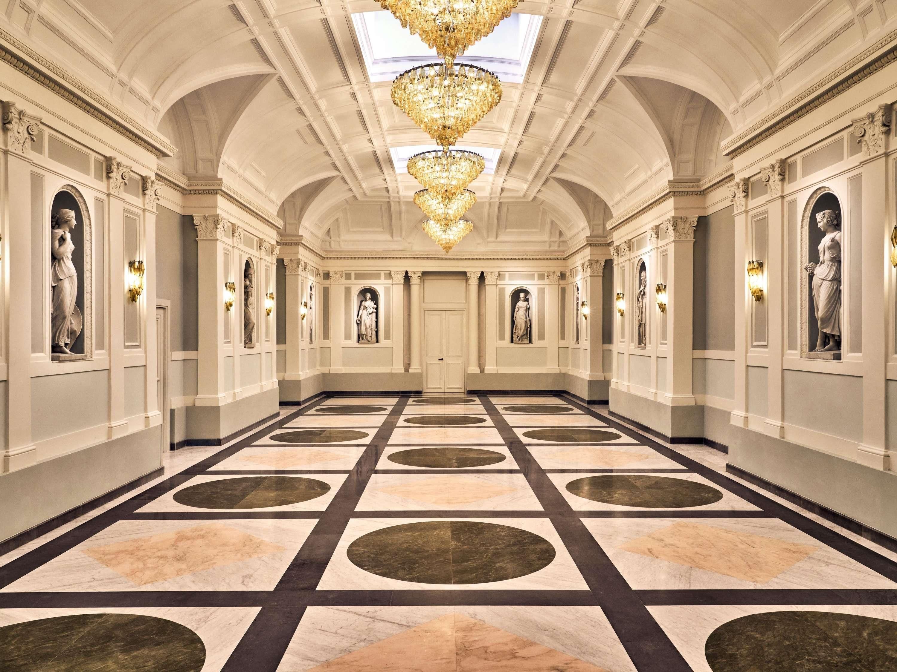 A grand hallway with a marble floor, classical statues, and ornate chandeliers.