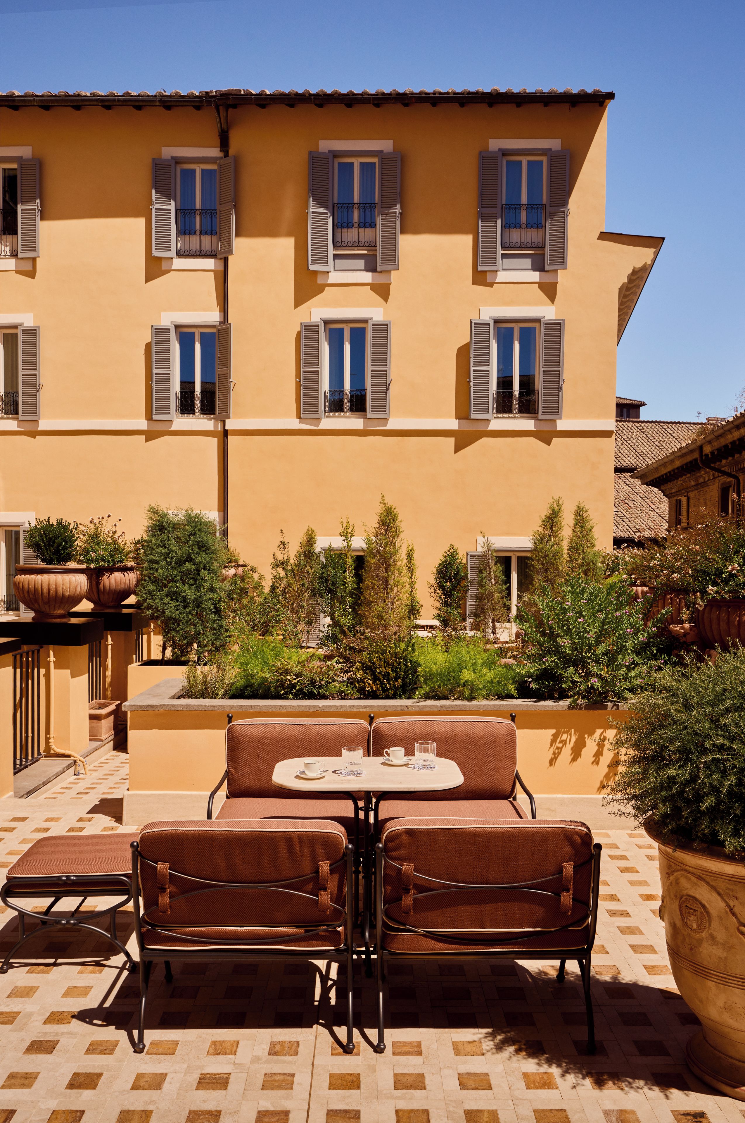 A terrace with a table and chairs, surrounded by potted plants and a building with multiple windows.