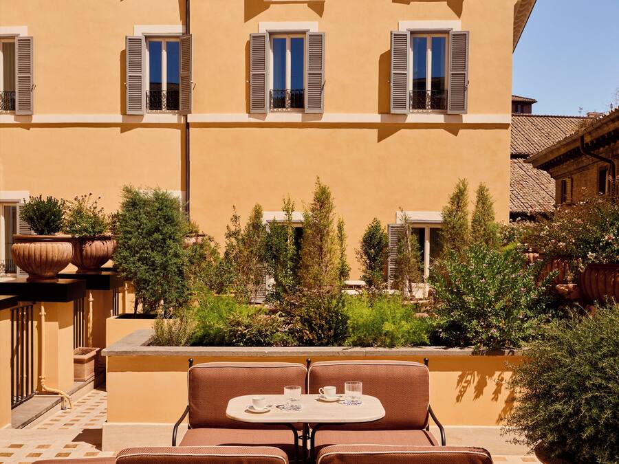 Una terraza con mesa y sillas, rodeada de macetas con plantas y un edificio con varias ventanas.