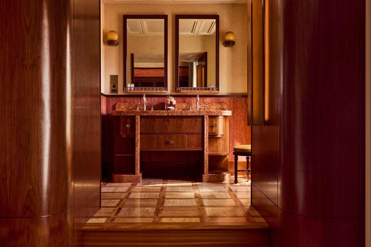 A wooden-floored hallway leading to a bathroom with a marble sink and double mirrors.