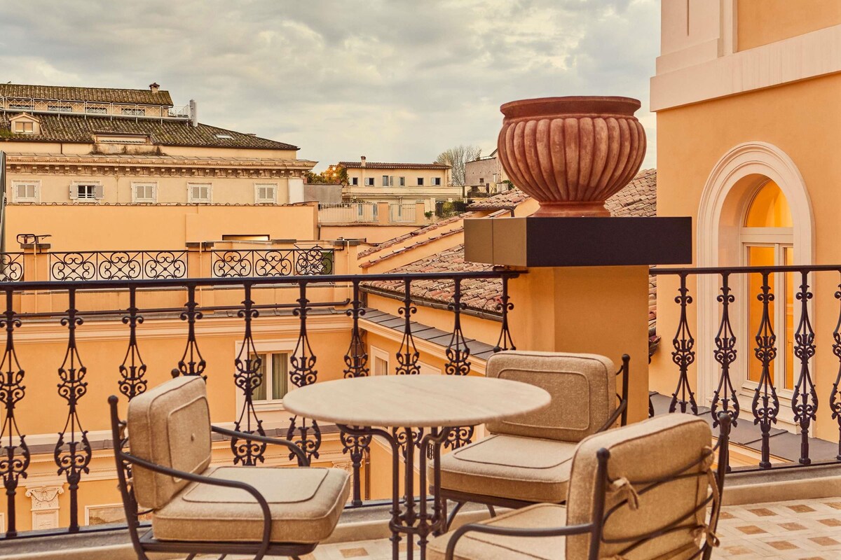 A balcony with a round table and two chairs, a large pot, and a view of surrounding buildings.