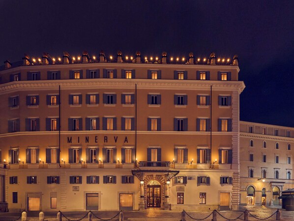 Un hôtel à plusieurs étages, dont la façade est ornée d’une enseigne bien visible portant le nom « MINERVA », et qui se caractérise par une disposition symétrique des fenêtres et une entrée centrale surmontée d’un auvent.