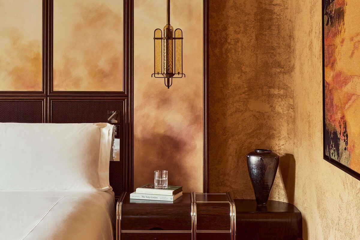 A hotel room with a bed, a bedside table with a book and a glass, a decorative vase, and a wall with a textured pattern.
