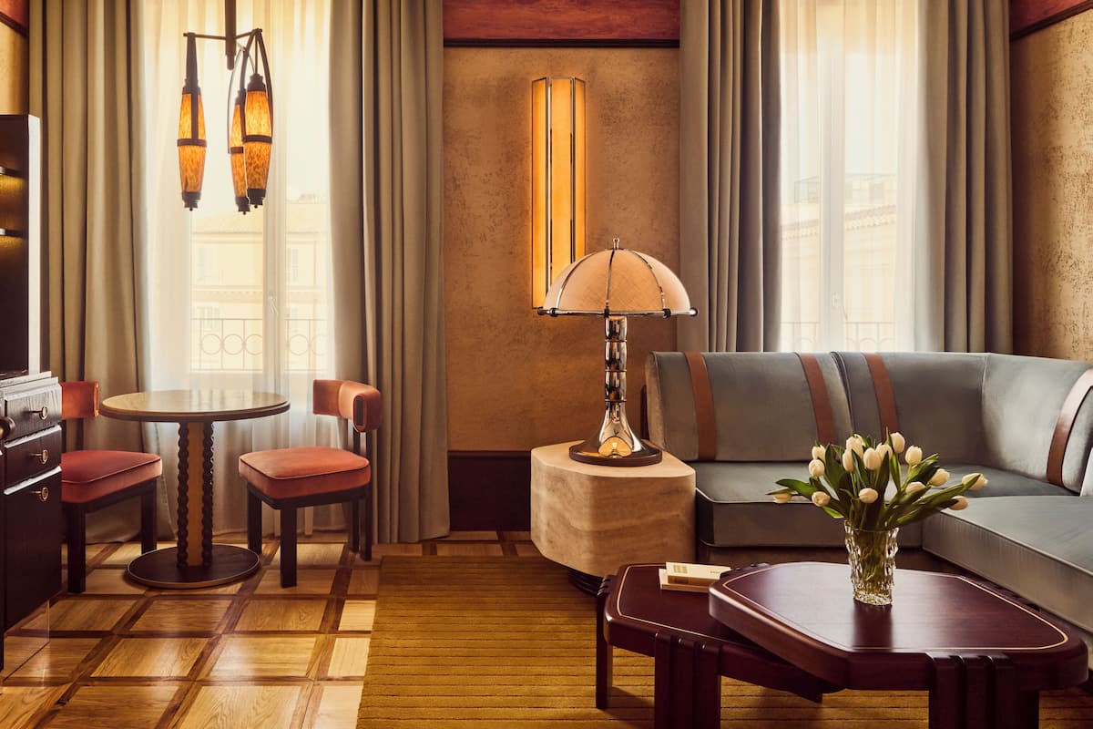 Une chambre d’hôtel avec un plancher en bois, une table ronde entourée de chaises, un canapé, une lampe et un vase rempli de fleurs.