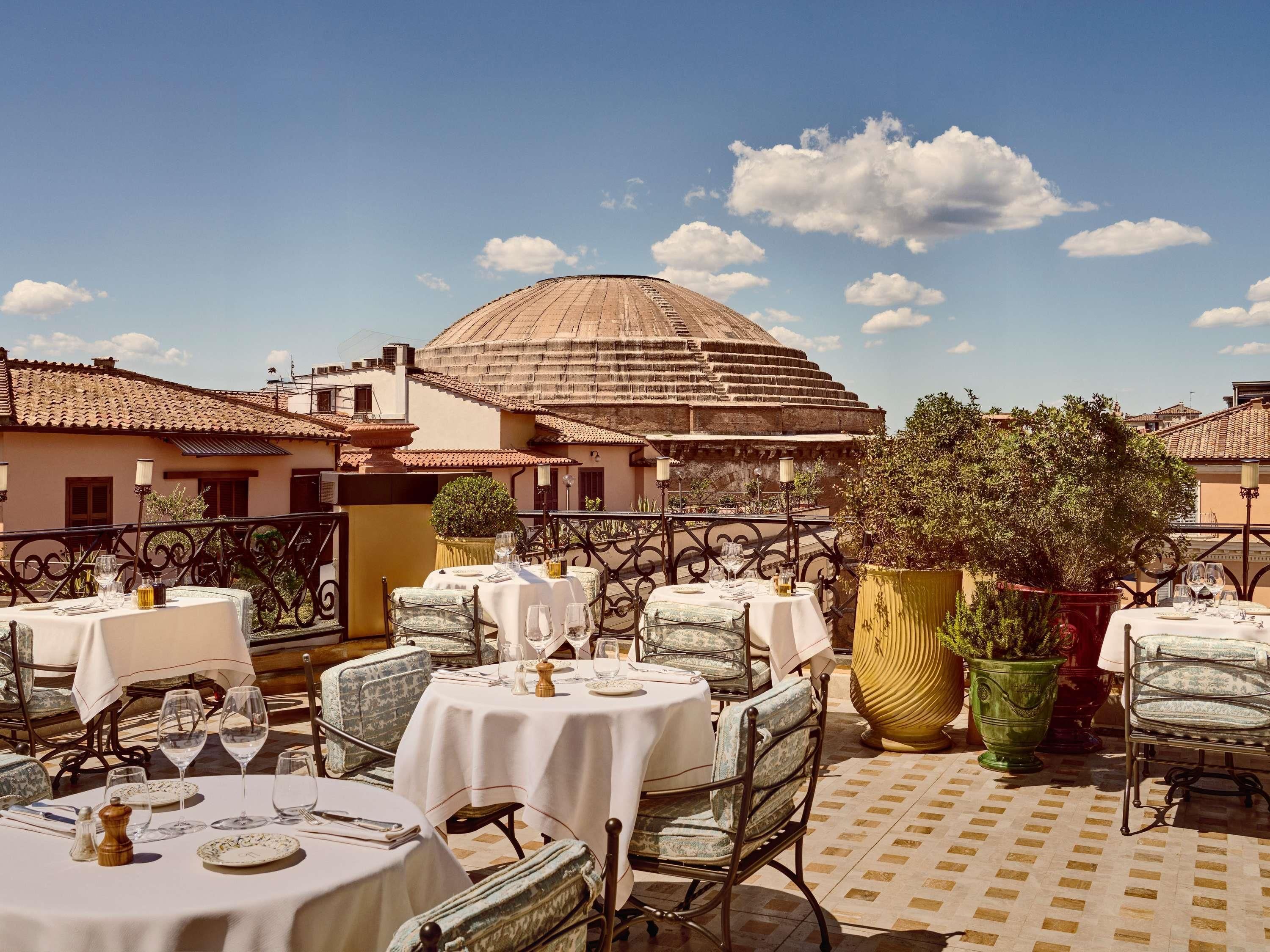 Un espace extérieur aménagé pour les repas, avec des tables et des chaises, une grande plante en pot et, au fond, un bâtiment surmonté d’un dôme.