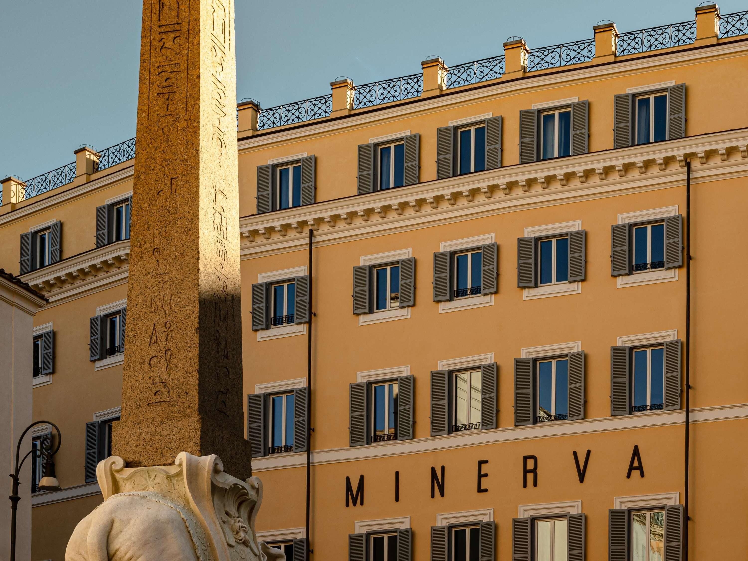 A building with the name 'MINERVA' on its facade, featuring multiple windows with shutters, a decorative column, and a wrought iron balcony railing.