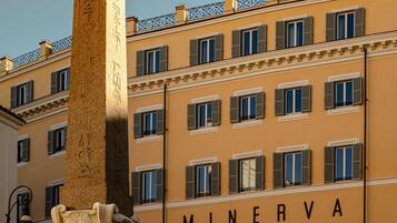 Un bâtiment dont la façade porte l’inscription « MINERVA », orné de nombreuses fenêtres avec volets, d’une colonne décorative et d’un garde-corps de balcon en fer forgé.