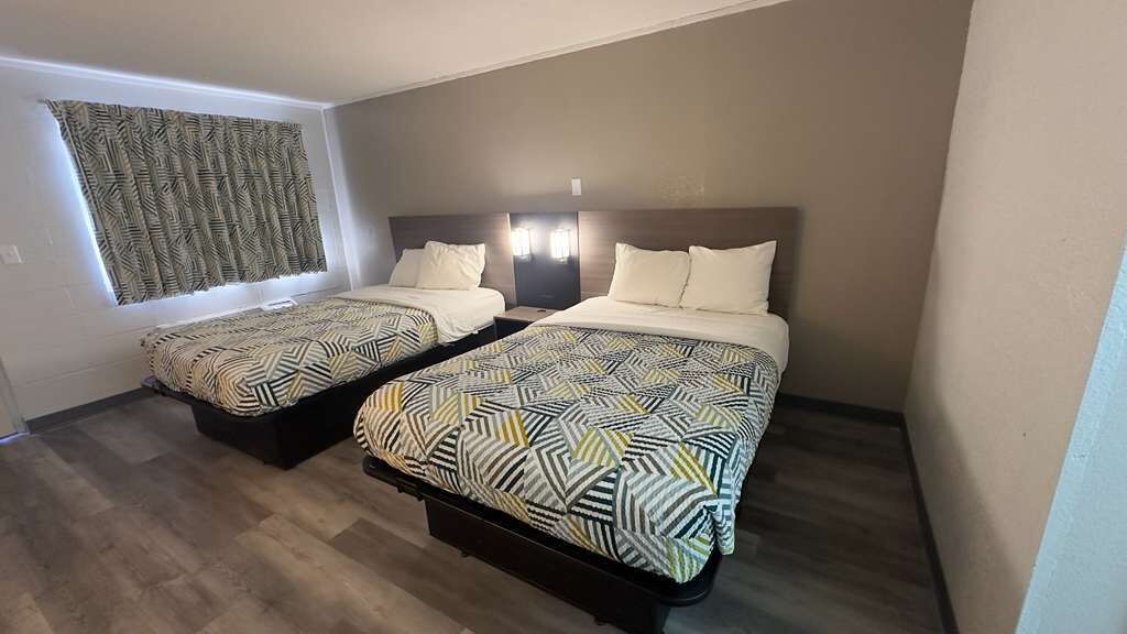 A hotel room with two beds, a window with patterned curtains, and a headboard with built-in lighting.