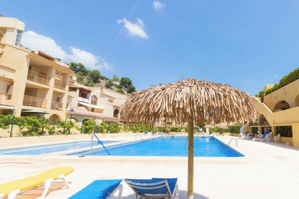A swimming pool with lounge chairs and a thatched umbrella, surrounded by buildings with balconies and greenery. - - BayAltea (Altea)